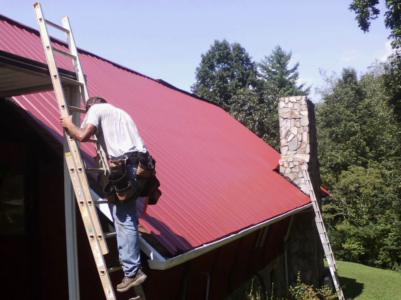 Roofer on a ladder beside a red metal roof and stone chimney — Soffit & Fascia Repair by Roofing Brothers in Beaver Falls